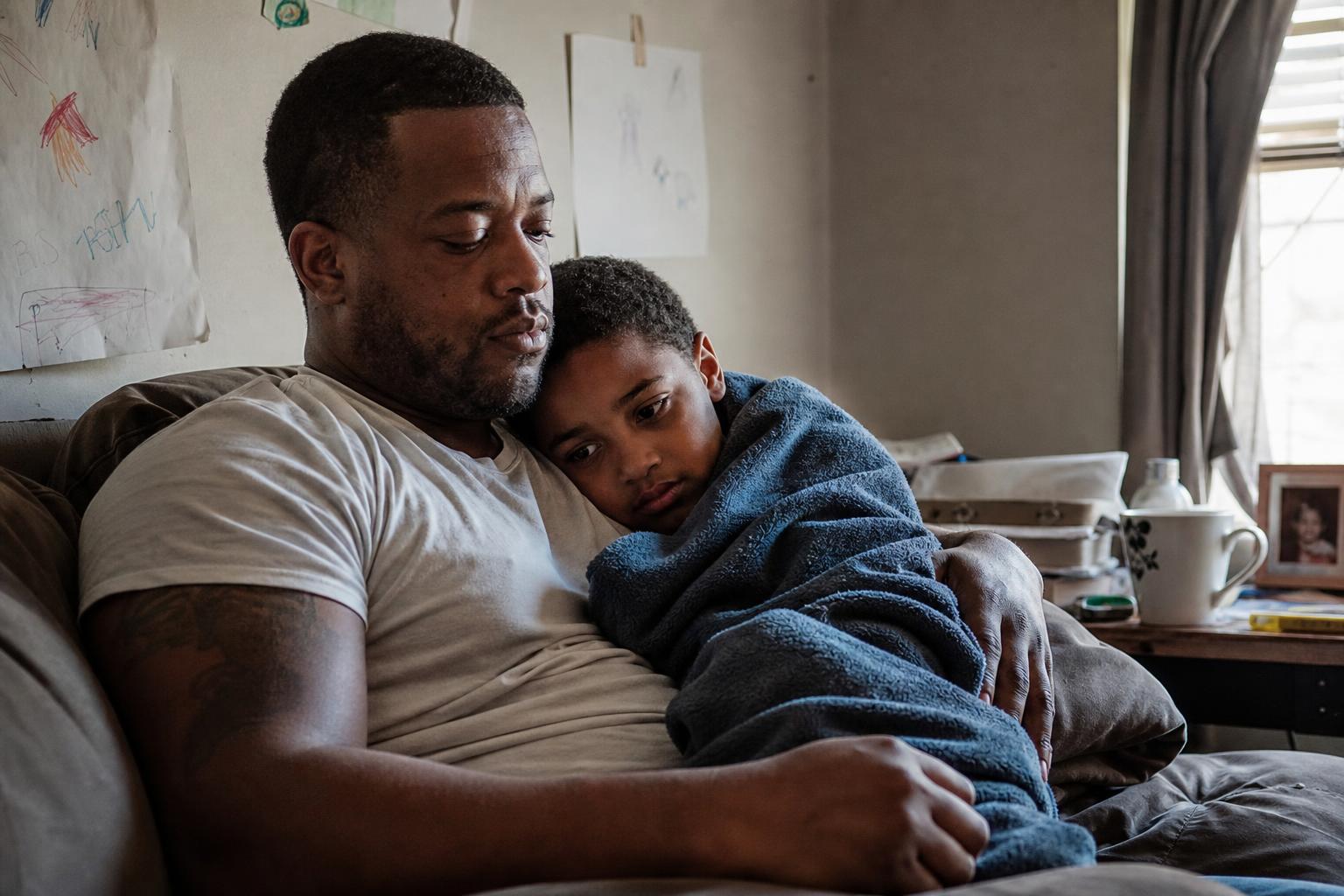 A young Black father comforting his school-age son on the couch in soft afternoon light.